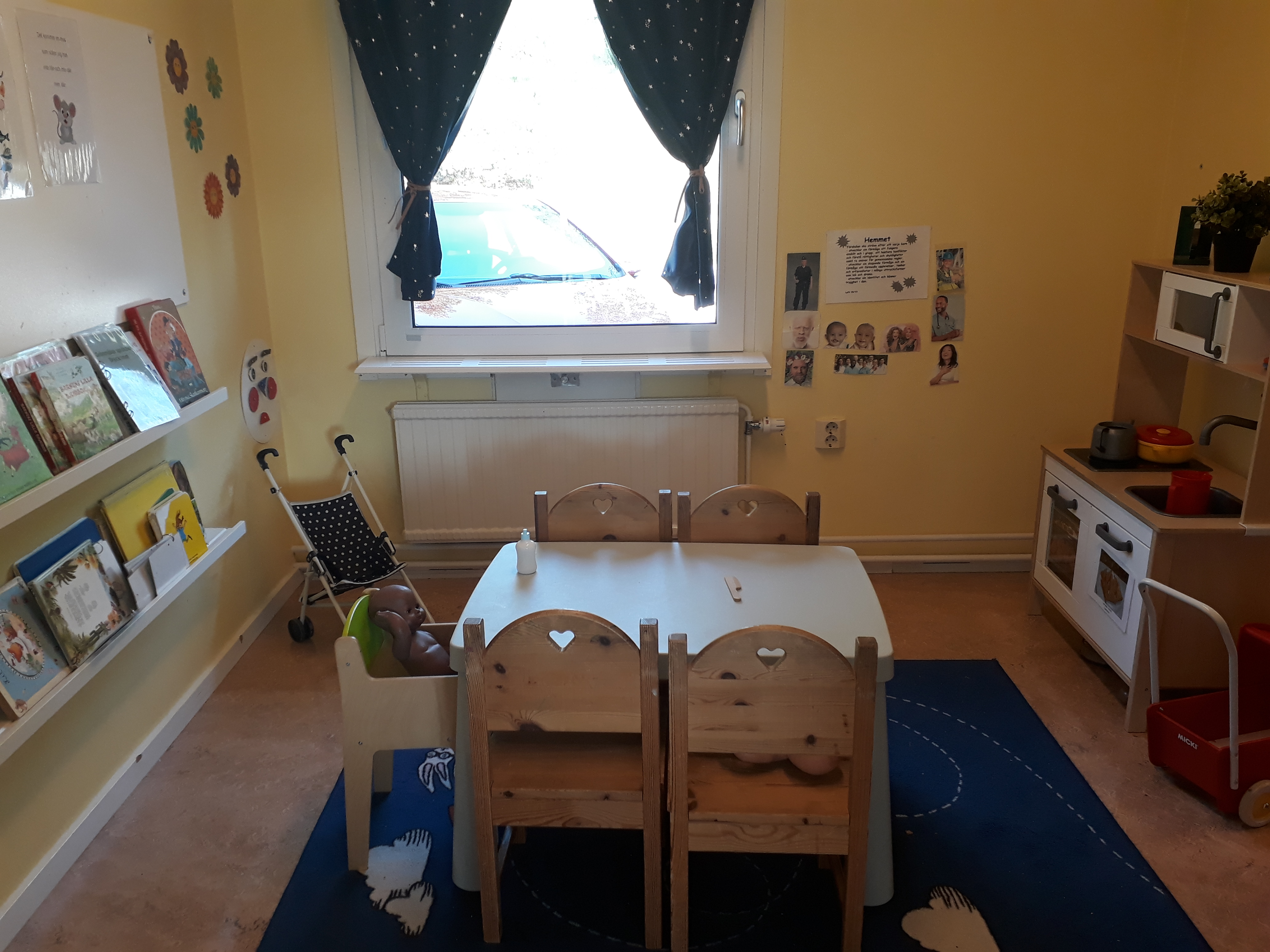 En leksaksspis står efter ena väggen. Ett fyrkantigt bord med stolar står på en blå matta mitt i rummet. En barnstol med en docka står också vid bordet. På väggen sitter en hylla med böcker.