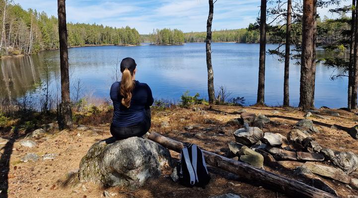 En kvinna sitter på en sten. I bakgrunden sjöutsikt över sjön Ötjärn.