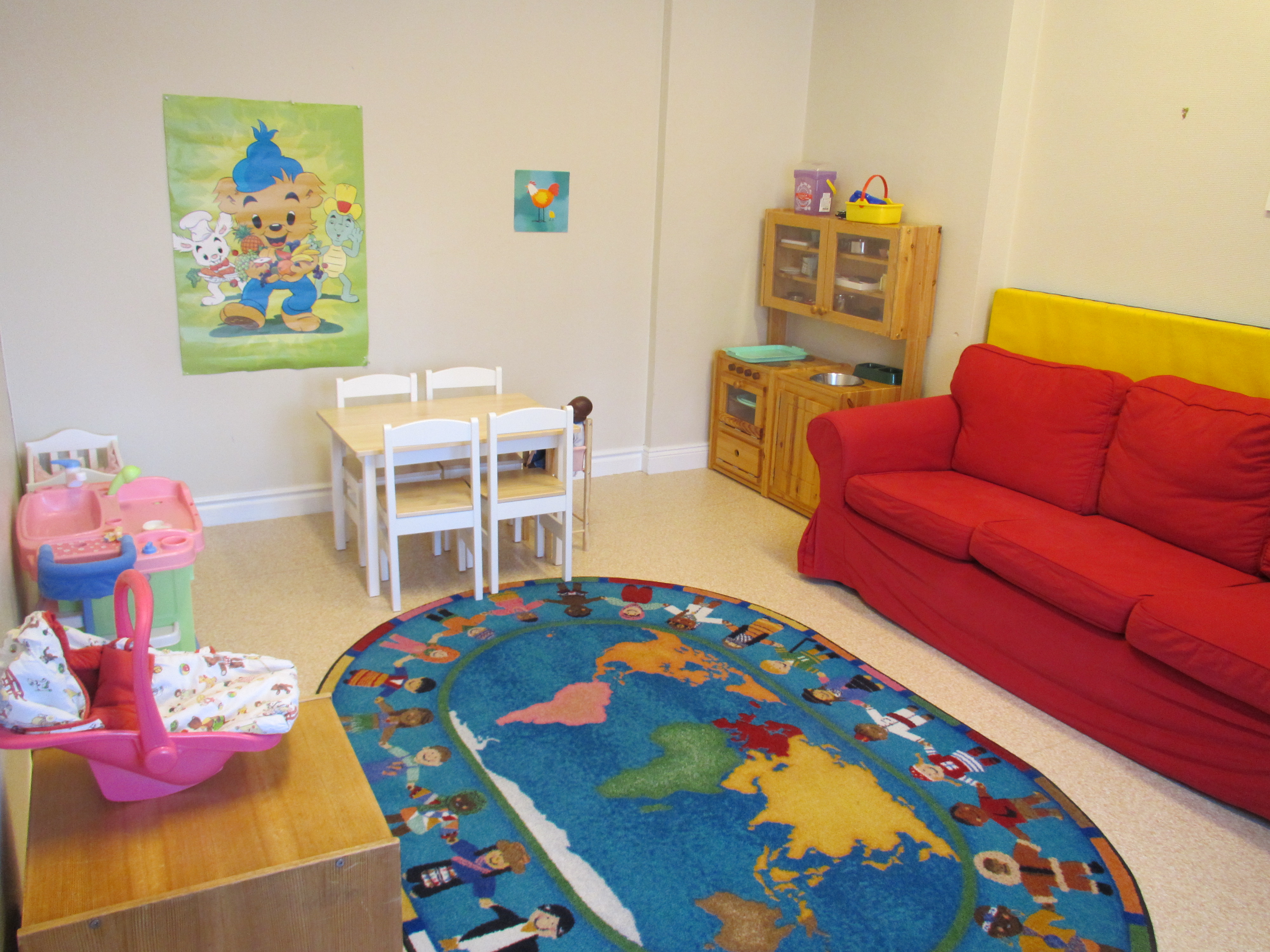 En stor färgglad matta ligger mitt på golvet. Efter en väggen står en röd soffa. Bredvid soffan står ett leksakskök. Ett litet fyrkantigt bord med stolar. En affisch på Bamse sitter på väggen.