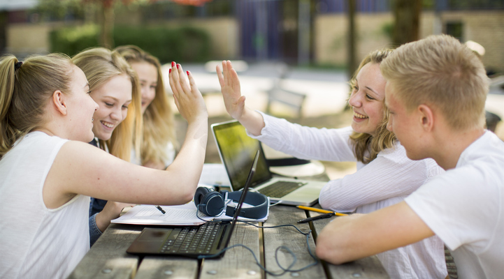 Fem elever sitter utomhus vid ett träbord. På bordet har dom laptops och papper. Två tjejer som sitter mitt emot varandra gör en high five.