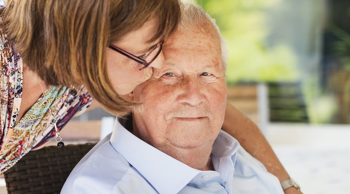 En kvinna pussar en äldre man på sidan om höger öga. Mannen sitter i en stol på en altan.