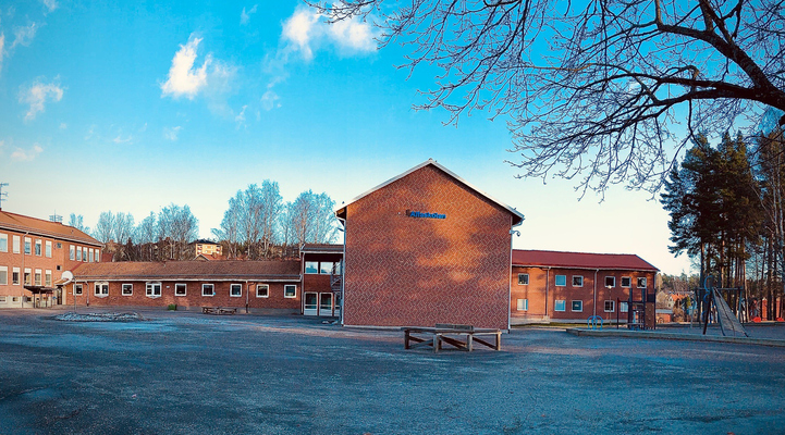Alfaskolans byggnad. En avlång röd tegelbyggnad med en blå skylt på där det står Alfaskolan. Lekpark syns till höger. Skolgård framför skolan.