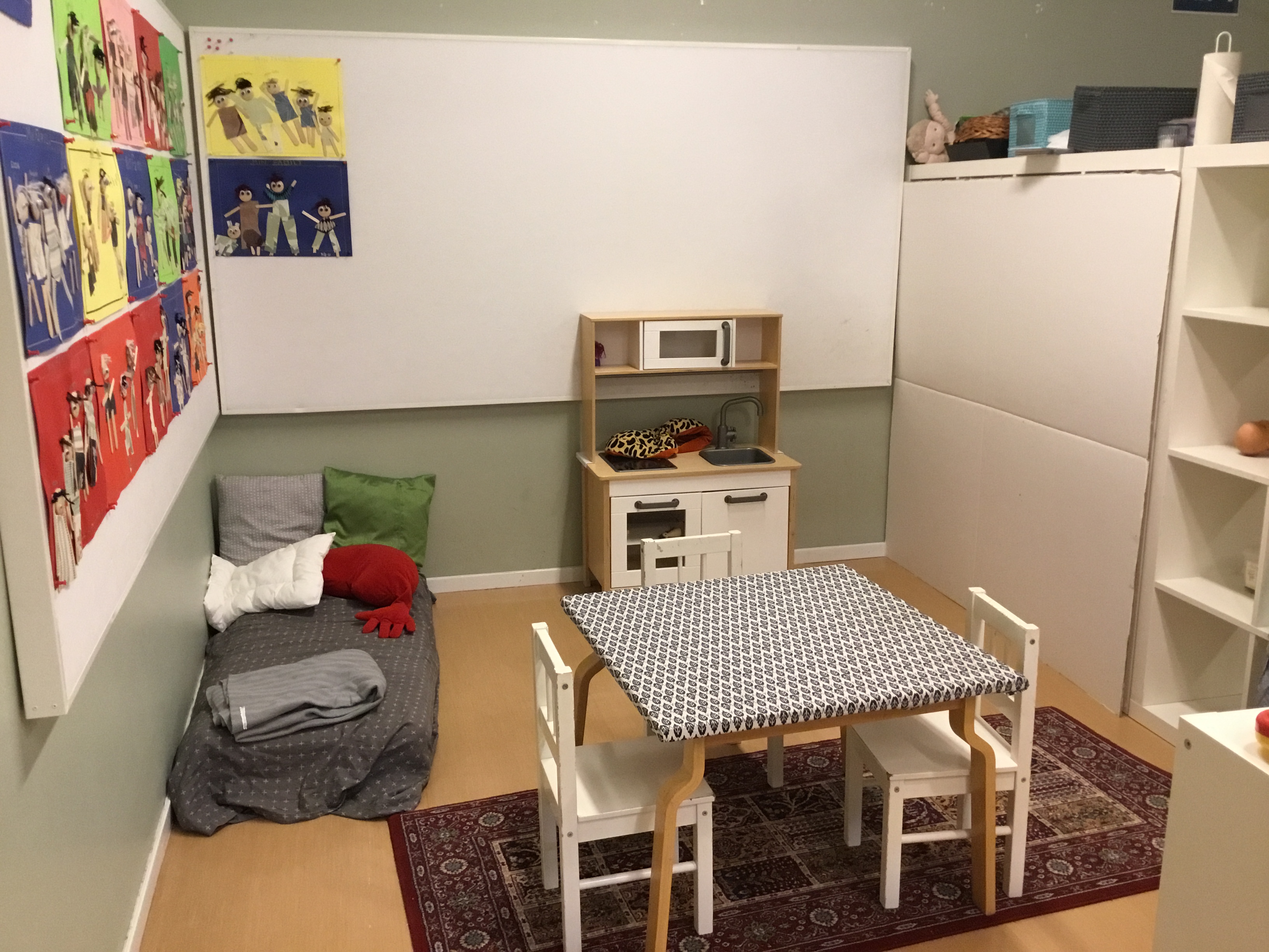 Ett leksaksspis står vid ena väggen, mitt i rummet står ett fyrkantigt bord och två stolar. En lite säng med grått täcke finns vid ena långväggen.