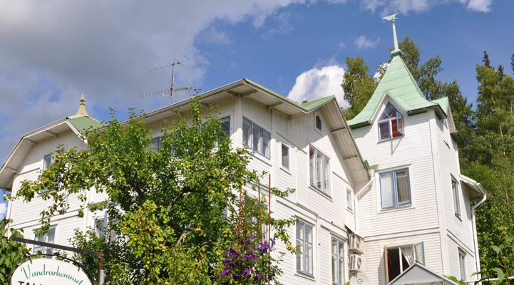 Vandrarhemmet Tallbacka i Ängelsberg. En vit villa med grönt plåttak. Grönska med träd omringar villan